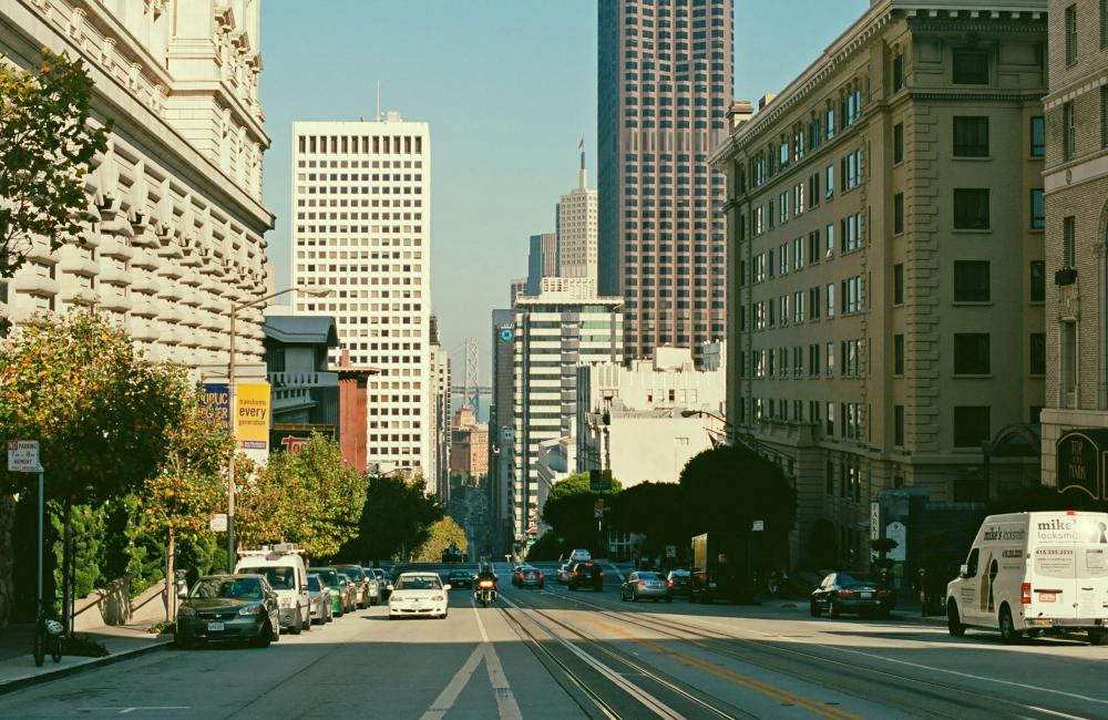 View of the middle of a street with tall buildings on both sides. The Bay Bridge can be seen between buildings.