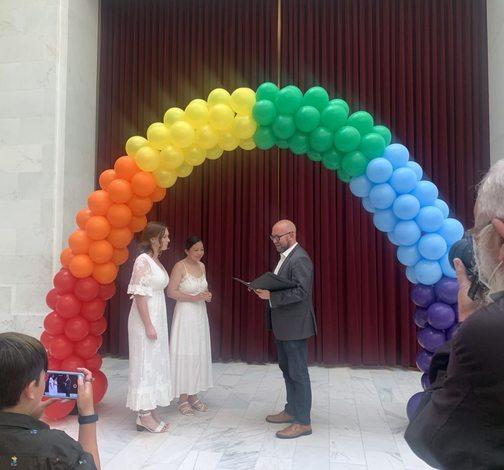 Supervisor Mandelman officiates a marriage of a couple both wearing white dresses in front of a rainbow balloon arch 
