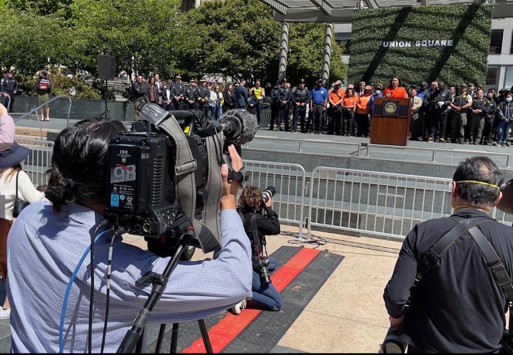 An SFGovTV camera operator captures a large press conference at Union Square, with Mayor London Breed at the lectern.