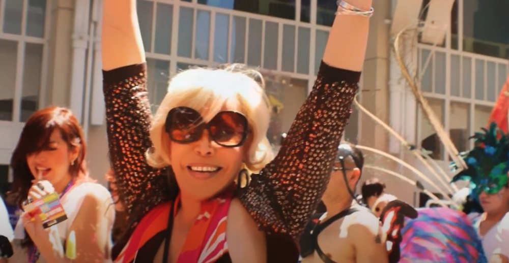 A person in a parade wearing sunglasses puts their hands up in the air to celebrate